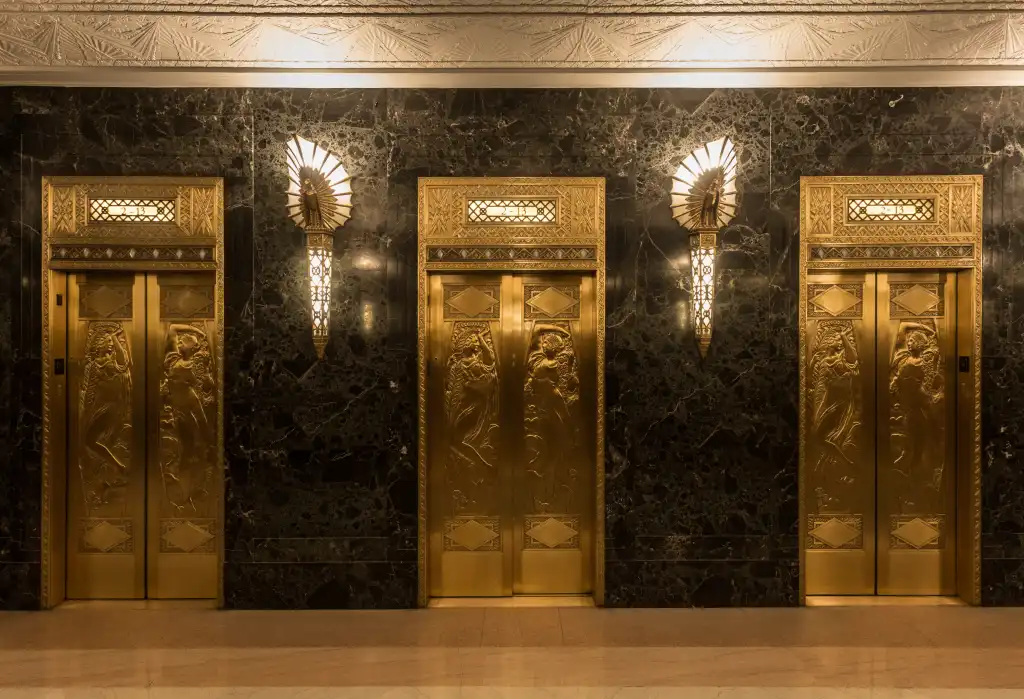 Dramatic golden elevators in fancy urban skyscraper building with relief sculptures and marble wall with lights and sconces