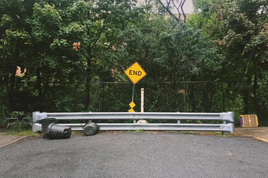 End of the road with End sign, barrier, tipped garbage cans, chain link fence, and green trees in the background