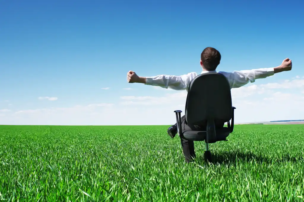 Young man with outstretched arms sitting in chair on green meadow and looking at a blue sky with clouds