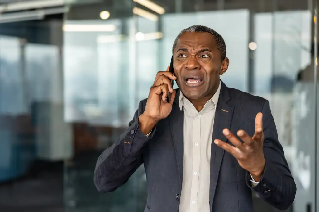 Mature businessman experiencing stress and frustration during a phone conversation, making an upset facial expression and gesturing with his hand in a modern office environment